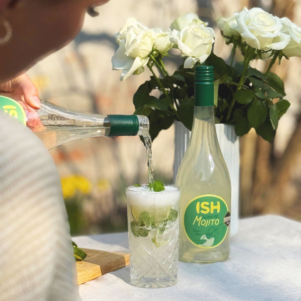 Person pouring a drink from a 'ISH Mojito' bottle into a glass with ice and mint leaves, surrounded by white roses.